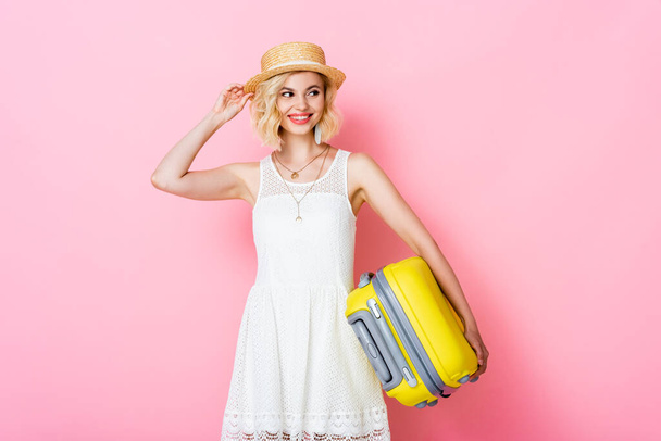 young woman touching straw hat while holding yellow luggage on pink  - Foto, Bild