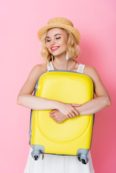  woman in straw hat embracing yellow luggage on pink  - 写真・画像