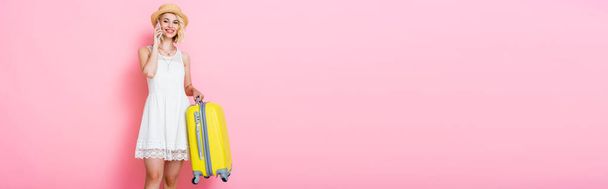 panoramiczna koncepcja kobiety w słomkowym kapeluszu trzymającej żółty bagaż i rozmawiającej na różowym smartfonie  - Zdjęcie, obraz