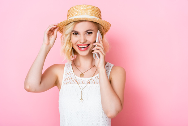 woman touching straw hat and talking on smartphone on pink  - Foto, imagen