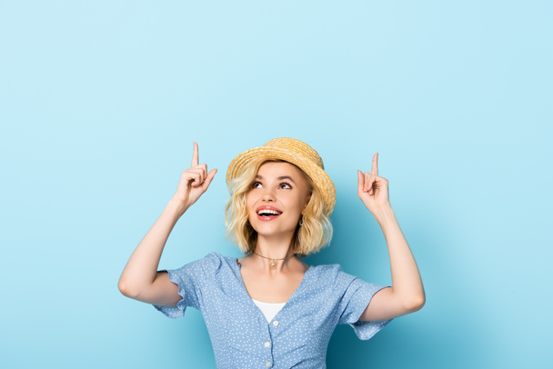 young woman in straw hat pointing with fingers and looking up on blue  - Foto, Imagem