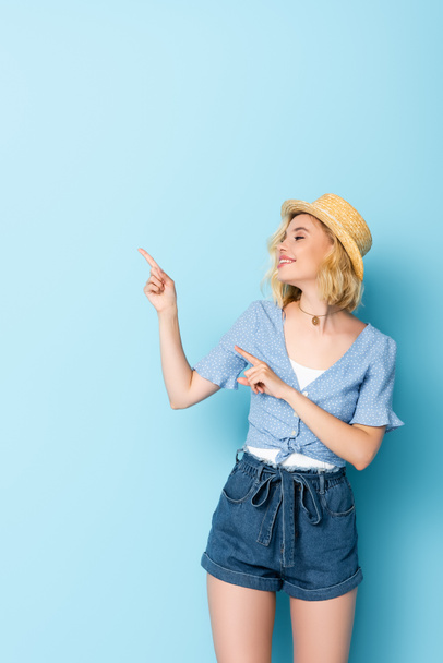 woman in straw hat pointing with fingers and looking away on blue  - Foto, immagini