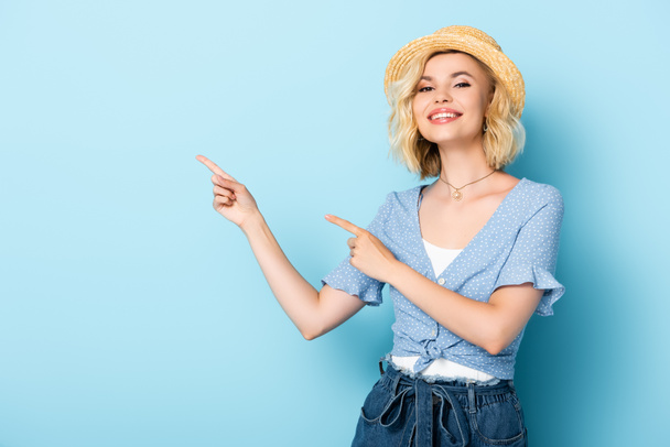 young woman in straw hat pointing with fingers and looking at camera on blue  - Foto, Imagen
