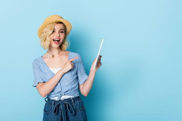 femme excitée en chapeau de paille et shorts pointant du doigt la tablette numérique sur bleu  - Photo, image