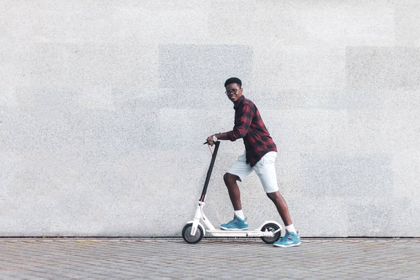 afro american guy rides an electric scooter against the background of a wall, a student uses eco transport - Foto, immagini