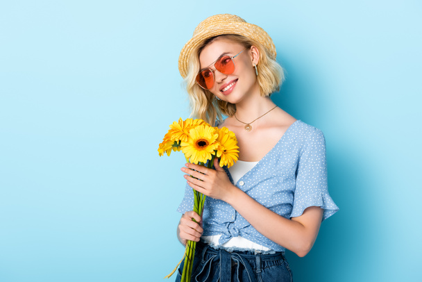 femme en chapeau de paille et lunettes de soleil tenant des fleurs sur bleu - Photo, image