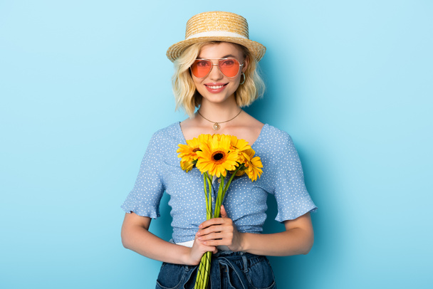 kobieta w słomkowym kapeluszu i okularach przeciwsłonecznych trzymających kwiaty na niebiesko - Zdjęcie, obraz