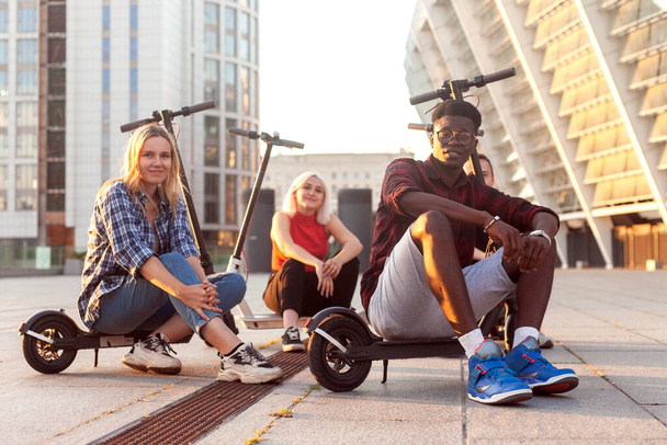 rotujenvälinen ryhmä nuoria ystäviä, jotka istuvat sähköskoottereilla kaupungissa ja hymyilevät, monirotuiset nuoret käyttävät ekokuljetuskonseptia - Valokuva, kuva