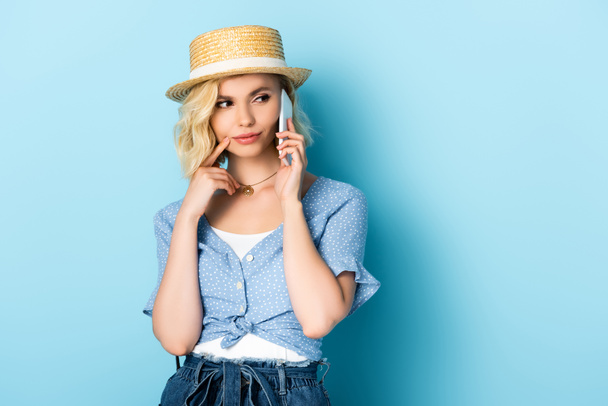 mujer en sombrero de paja mirando hacia otro lado y hablando en el teléfono inteligente en azul  - Foto, imagen