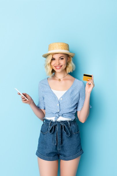 young woman in straw hat holding credit card and smartphone on blue  - Foto, imagen