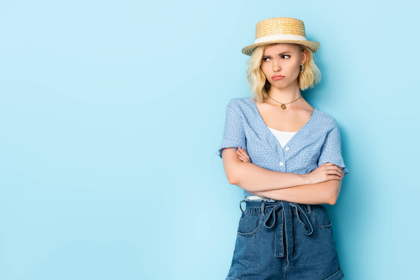 niezadowolona młoda kobieta w słomkowym kapeluszu stojąca z skrzyżowanymi ramionami i odwracająca wzrok na niebiesko  - Zdjęcie, obraz