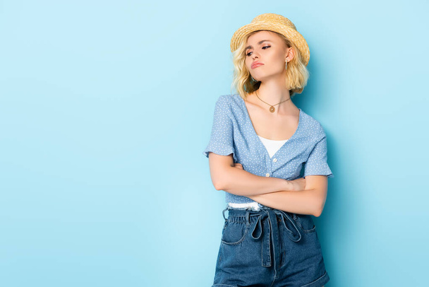 offended woman in straw hat standing with crossed arms and looking away on blue  - Foto, Imagen