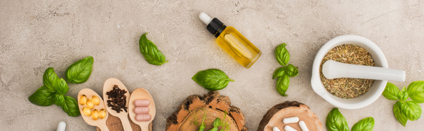 panoramic shot of herbs, green leaves, mortar with pestle, bottle and pills in wooden spoons on concrete background, naturopathy concept - Photo, Image