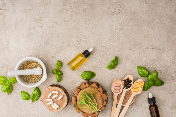 top view of herbs, green leaves, mortar with pestle, bottles and pills in wooden spoons on concrete background, naturopathy concept - Photo, Image