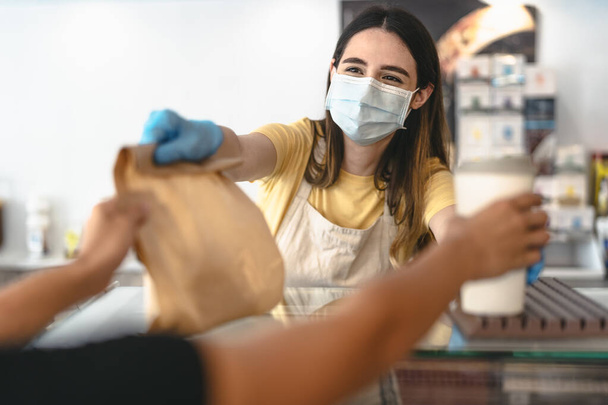 Bar owner working only with take away orders during corona virus outbreak - Young woman worker wearing face surgical mask giving takeout meal to customers - Healthcare and Food drink concept - Photo, image