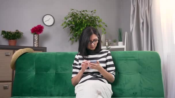 Charming serious modern young woman in glasses and in stylish wear sitting on comfortable sofa at home and using her smartphone - Séquence, vidéo