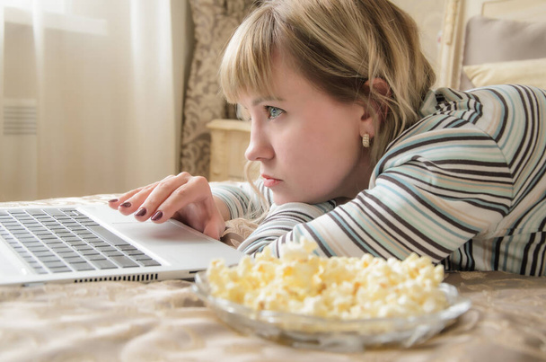 A young blonde woman lies and looks intently into the laptop screen. Close-up portrait. Concept: watching movies online, leisure at home, communicating in social networks. - Photo, image
