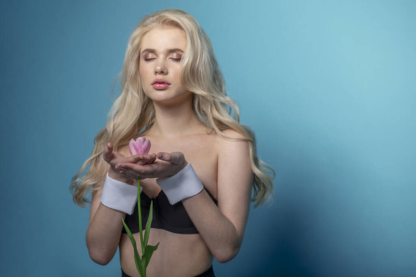 Приваблива блондинка молода жінка позує в студії, тримаючи рожевий тюльпан в руках, одягнена в чорну білизну на синьому фоні
. - Фото, зображення