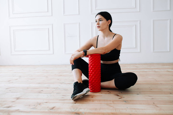 Young Asian woman in a black sports top and leggings sits on a wooden floor with a massage roller and rests after a workout in the classic interior - Φωτογραφία, εικόνα