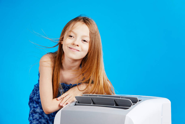 cute smiling little caucasian girl wearing a summer dress next to an air conditioning on a blue studio background - Φωτογραφία, εικόνα