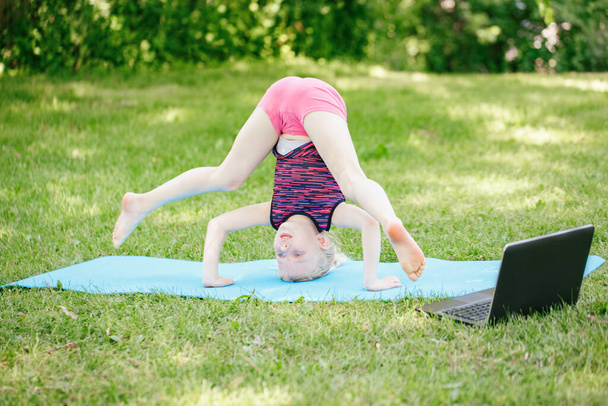 Dívka dítě dělá sportovní cvičení venku online. Video jóga protahování na internetu v parku. Dítě učí fitness gymnastiku na dvorku s laptopem. Nový normální. Společenská vzdálenost u koronaviru. - Fotografie, Obrázek