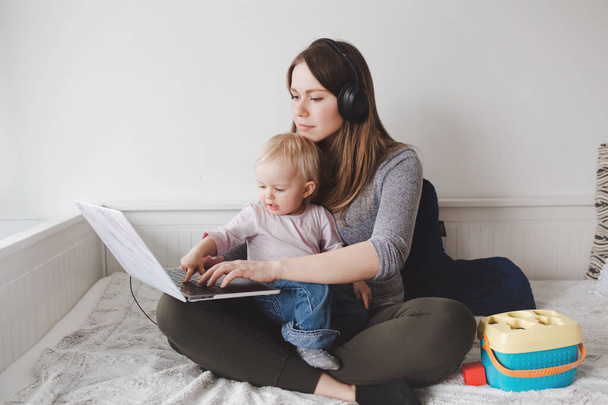 Caucasian mother blogger with baby working on laptop from home. Workplace of freelance woman with kid toddler. Stay at home single mom earning money at online remote distant job. - Photo, Image