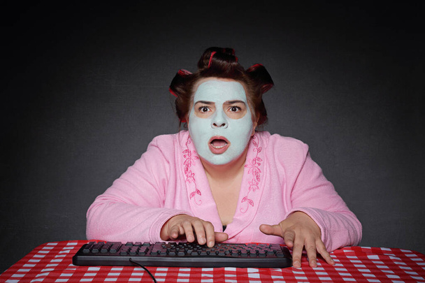 lustige übergewichtige fünfzigjährige Frau mit Bademantel und Gesichtsmaske frustriert auf abhörsicherem Laptop auf Studiograuen Hintergrund - Foto, Bild