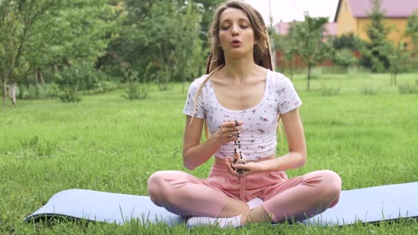 jong mooi meisje met lange dreadlocks houdt in handen de ketting gemaakt door natuurlijke stenen voor meditatie buiten tijdens de ochtend op groen gras van haar achtertuin - Video
