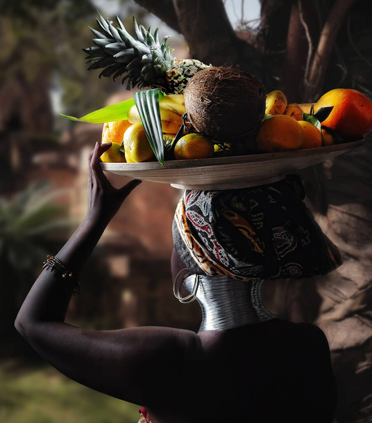 travel photography, african black woman portrait in ethnic dress rear view with fruit dish on her head on tropical background - Photo, Image