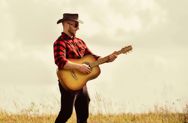 Inspired country musician. Country style. Summer vacation. Country music concept. Guitarist country singer stand in field sky background. Hiking song. Play beautiful melody. Handsome man with guitar - Photo, image