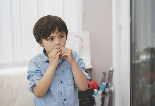 Portret van een kind dat op zijn vingernagels bijt terwijl hij naar iets kijkt, emotioneel kinderportret dat alleen staat met een denkend gezicht, jonge jongen die vinger in zijn mond steekt. - Foto, afbeelding