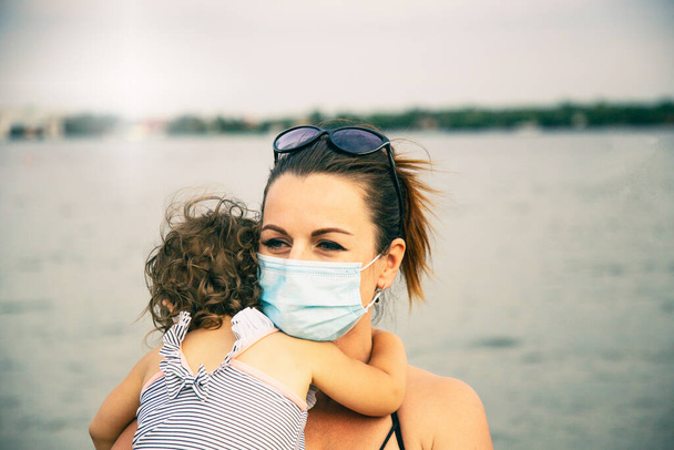 Kaukasisches Kleinkind und seine Mutter tragen Schutzmasken am Sandstrand. Sommerurlaub nach der Coronavirus-Pandemie. - Foto, Bild