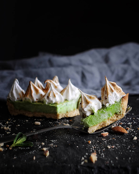 food photography of sliced sweet homemade dessert tart side view close up with green cream white toasted meringue on a dark textural background with a fork, mint and nuts - Foto, afbeelding