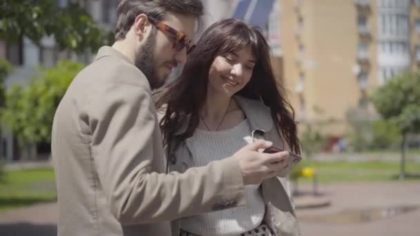Bearded Caucasian man and smiling brunette woman using smartphone as meeting friend on sunny city street. Portrait of positive friends greeting each other and leaving. - Кадры, видео