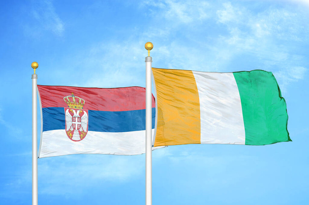 Serbia y Costa de Marfil Costa de Marfil dos banderas sobre asta de bandera y fondo azul cielo nublado - Foto, Imagen