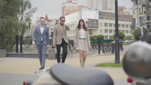 Portrait of three positive male and female biker in elegant clothes coming to scooters standing on sunny summer city street. Adult Caucasian group of friends resting outdoors. - Πλάνα, βίντεο