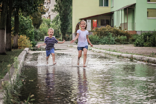 Cute happy kids jumping in the puddles after warm summer rain, outdoor walking w każdą pogodę, szczęśliwe dzieciństwo i lato, lifestyle outdoor, widok z tyłu - Zdjęcie, obraz