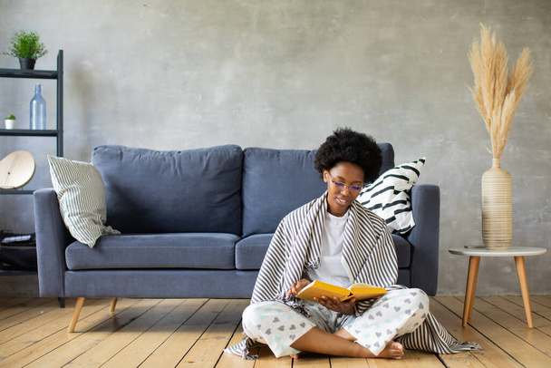 Szczęśliwa Afroamerykanka siedzi na wygodnej kanapie i czyta książkę. Szczęśliwy, aby przenieść się do nowego mieszkania. - Zdjęcie, obraz