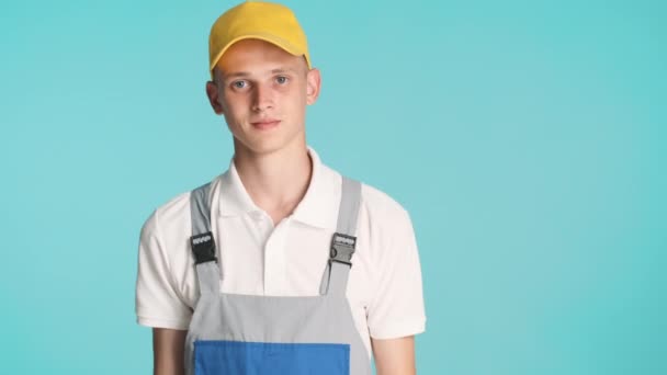 Jeune constructeur attrayant en uniforme et chapeau jaune montrant pouce vers le haut sur la caméra sur fond coloré - Séquence, vidéo