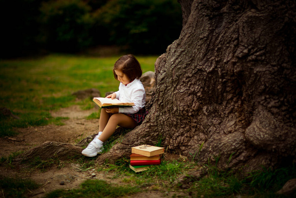una niña en uniforme escolar, comiendo un sándwich cerca de un árbol grande, durante un descanso entre clases en la escuela, 1 de septiembre - Foto, Imagen