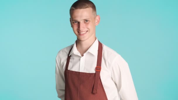 Młody przystojny, pewny siebie kelner w mundurze uśmiechnięty przed kamerą na niebieskim tle - Materiał filmowy, wideo