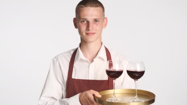 jonge knappe ober in schort houden lade met wijn zelfverzekerd kijken in camera over witte achtergrond - Video