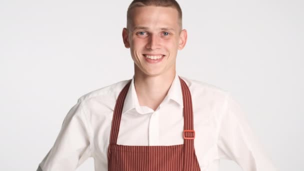 Młody przystojny wesoły kelner w mundurze uśmiechnięty do kamery na białym tle. Szczęśliwa ekspresja - Materiał filmowy, wideo