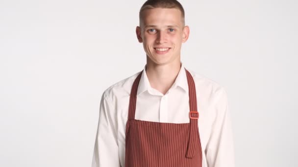 Young handsome waiter in uniform happily looking in camera over white background - Footage, Video