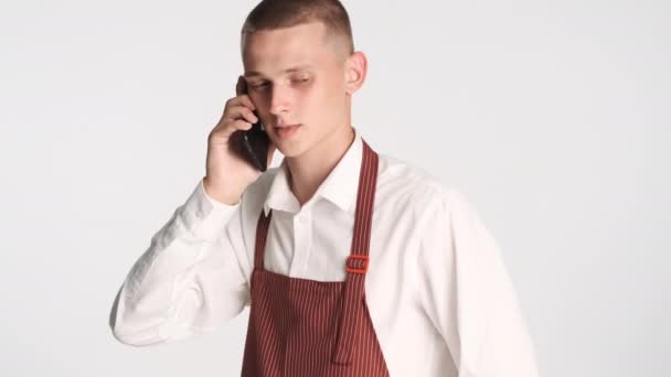 Junge attraktive Kellnerin in Uniform hört auf, auf dem Smartphone vor weißem Hintergrund zu reden - Filmmaterial, Video
