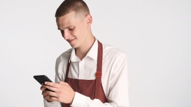 Joven camarero atractivo en uniforme felizmente usando teléfono inteligente sobre fondo blanco - Metraje, vídeo