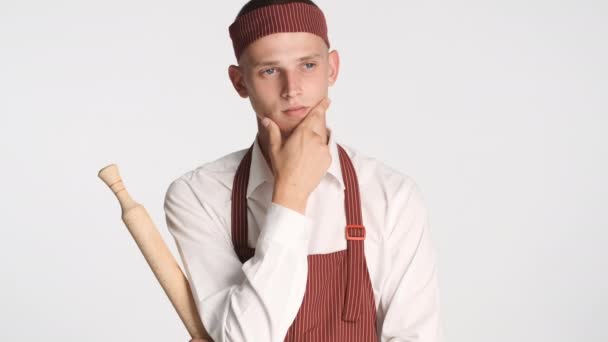 Fiatal vonzó szakács szakács egyenruhában gördülő pin figyelmesen pózol a kamera felett fehér háttér. Gondolkodó kifejezés - Felvétel, videó