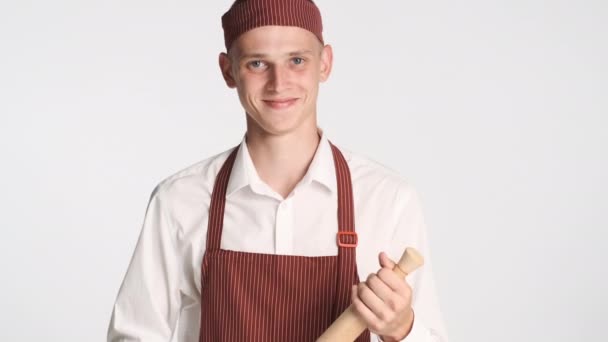Joven cocinero atractivo en uniforme con rodillo sonriendo alegremente en la cámara sobre fondo blanco - Imágenes, Vídeo
