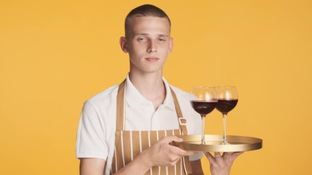 Joven camarero seguro atractivo en delantal sosteniendo bandeja con vino sonriendo en la cámara sobre fondo colorido - Metraje, vídeo