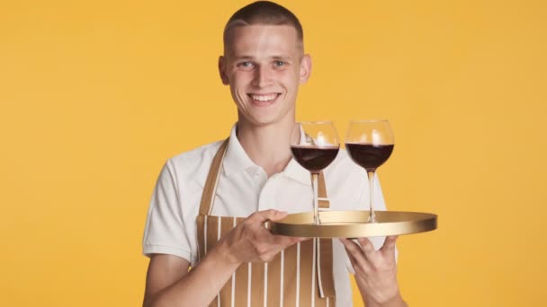 Fiatal vonzó vidám pincér kötényben tartó tálca bor boldogan néz kamerába színes háttér - Felvétel, videó